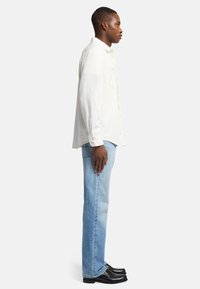 Camicia bianca a maniche lunghe con dettagli a bottoni, abbinata a jeans blu chiaro a gamba dritta e scarpe in pelle nere, vista di lato.