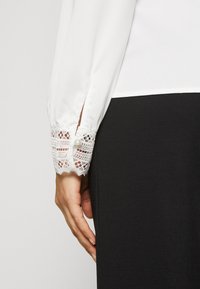 Blouse blanche avec bande de dentelle aux poignets, ornée d'un bouton décoratif ; associée à un vêtement noir en dessous.