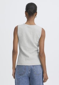 Frau mit geflochtenem Dutt, die silberne Creolen trägt, ein ärmelloses graues Oberteil und blaue Jeans aus Denim, stehend mit dem Rücken zur Kamera.