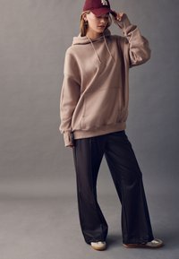 Beżowa, oversize’owa bluza z kapturem i kieszenią typu kangur, zestawiona z czarnymi szerokimi spodniami i beżowymi sneakersami. Burgundowa czapka z białym logo.