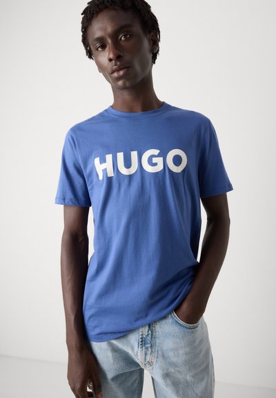 Синя памучна тениска с кръгло деколте, сbold бел текст "HUGO" на гърдите. Прост дизайн, къси ръкави, мека текстура.