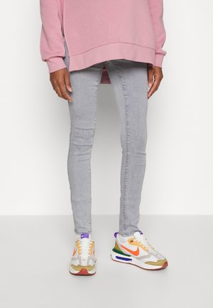 Personne portant un jean skinny gris clair, des baskets blanches et orange, et un sweat-shirt rose oversize, debout sur un fond uni.