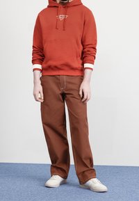 Röd hoodie med vitt logotyp, kenguruficka och vita randiga manschetter. Bruna vida byxor med kontrasterande sömmar. Vita sneakers.