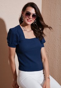 Navyblaues Top mit quadratischem Ausschnitt und kurzen Puffärmeln, aus strukturierter Stoff, kombiniert mit weißen eng anliegenden Hosen und silbernem Schmuck.