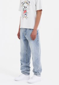 Jeans de lavado claro, de corte relajado, con un logo bordado en el bolsillo trasero, combinados con una camiseta gráfica gris y zapatillas blancas.