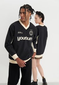 Top negro de manga larga con escote en V, detalles en crema y el logo "yourturn". El modelo lo lleva puesto con pantalones negros, con un fondo texturizado visible.