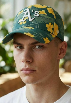 Young man with light skin wearing a green and yellow floral baseball cap with a white "A's" logo, looking directly at the camera.