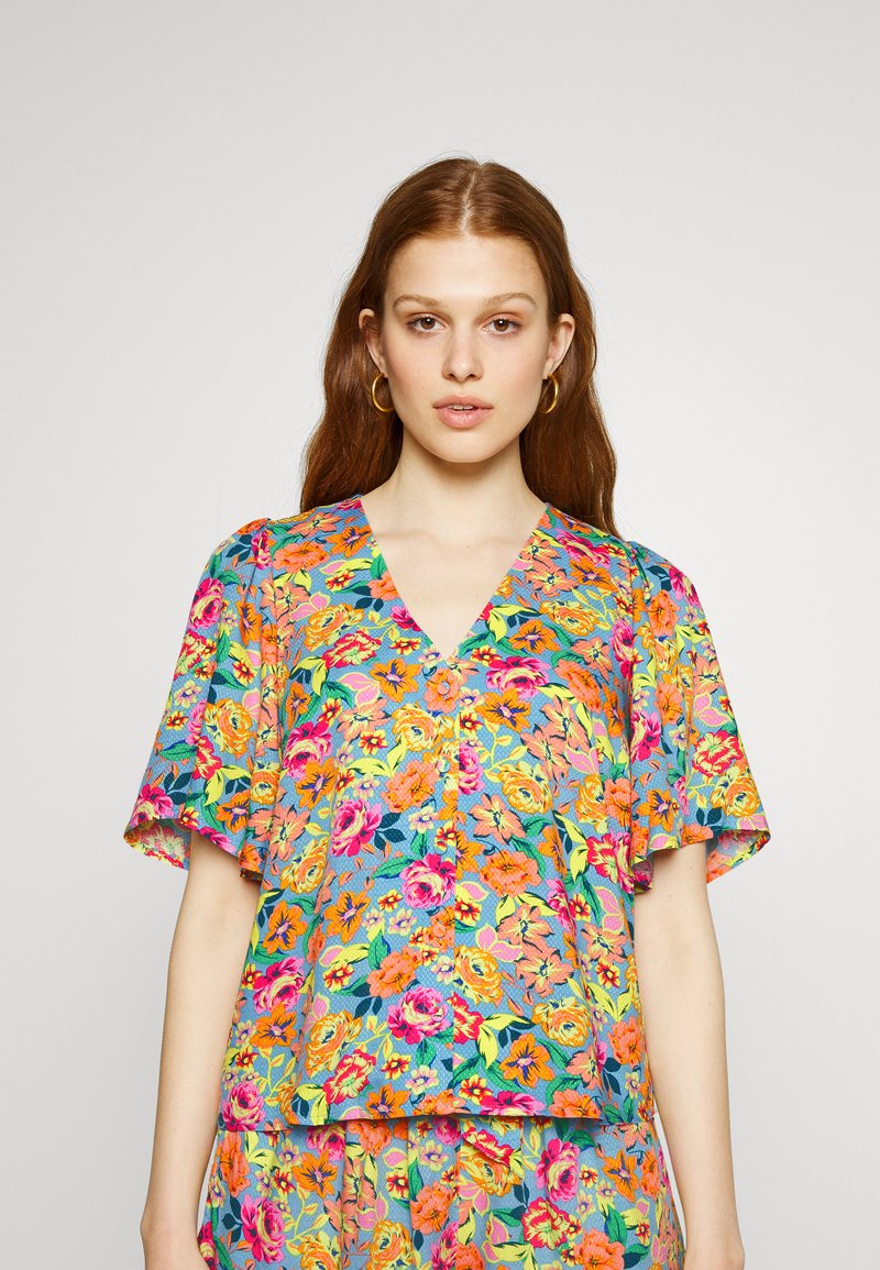 Blusa floral com mangas curtas, apresentando um fundo azul e flores vibrantes de várias cores. Design de decote em V com um ajuste relaxado.