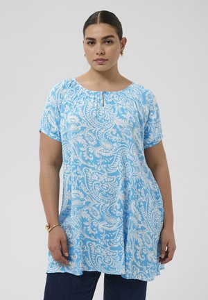 Nő világoskék tunikát visel fehér paisley mintákkal, rövid ujjakkal és kerek nyakkivágással, sötét nadrággal és arany karika fülbevalóval párosítva.
