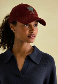 Bordeauxfarbene Corduroy-Mütze mit gesticktem Design auf der Vorderseite. Kombiniert mit einem marineblauen Pullover mit V-Ausschnitt und strukturierten Strickdetails.