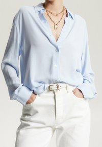 Camicia azzurro chiaro con maniche lunghe, realizzata in tessuto morbido. Abbinata a pantaloni bianchi a vita alta e a una cintura bianca con accessori dorati.