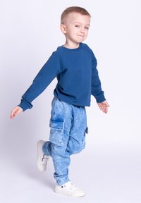 Niebieska koszula z długim rękawem, niebieskie spodnie typu cargo z efektem tie-dye i bocznymi kieszeniami oraz białe trampki. Casualowy strój o kontrastujących kolorach i luźnym kroju.