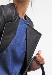 Veste en cuir noir avec des épaules texturées, associée à un haut en denim bleu avec une poche. Le modèle porte un sac à bandoulière noir.