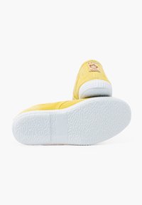 Zapatillas de tela amarillas con una suela de goma blanca. Cuentan con un parche en el talón y una suela texturizada para mayor agarre. Diseño simple y de perfil bajo.