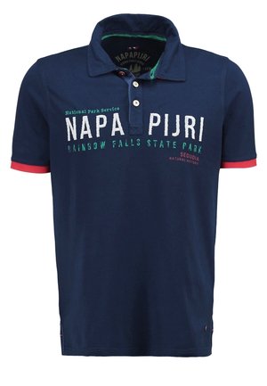 Mann trägt ein marineblaues Polohemd mit Schriftzug, beige Slim-Fit-Jeans und braune Sneaker, steht mit einer Hand in der Tasche.
