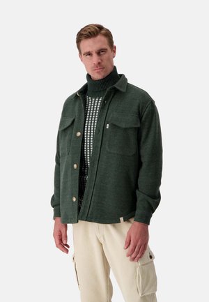 Grüne Wollmischung-Hemdjacke mit zwei Brusttaschen und beigen Knöpfen, über einem gemusterten Rollkragenpullover getragen und kombiniert mit cremefarbener Cordhose.