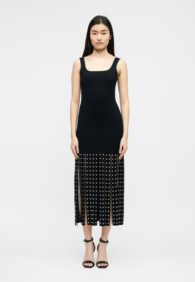 Γυναίκα που φοράει μαύρο midi φόρεμα χωρίς μανίκια με εφαρμοστό μπούστο και πλισέ φούστα με κυκλικές διακοσμήσεις, σε συνδυασμό με μαύρες γόβες με λουράκι στον αστράγαλο.