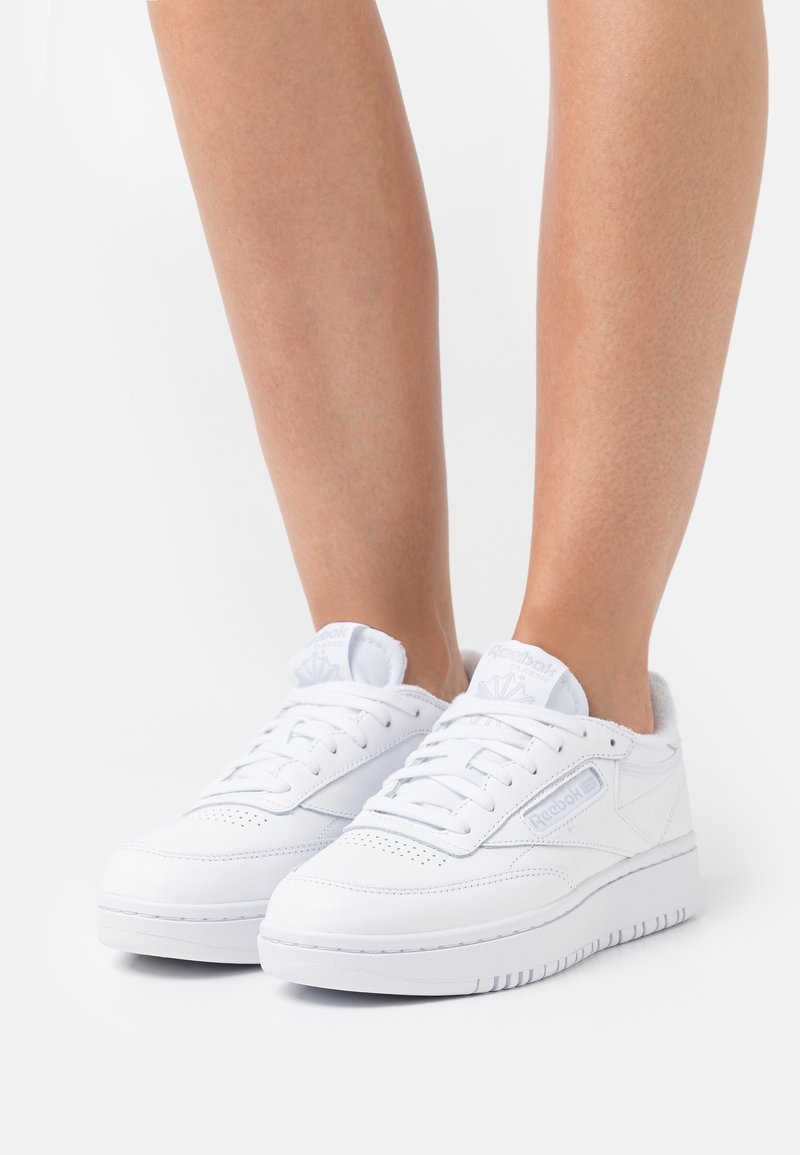 Personne portant des baskets en cuir blanches Reebok avec lacets, debout contre un fond clair uni.