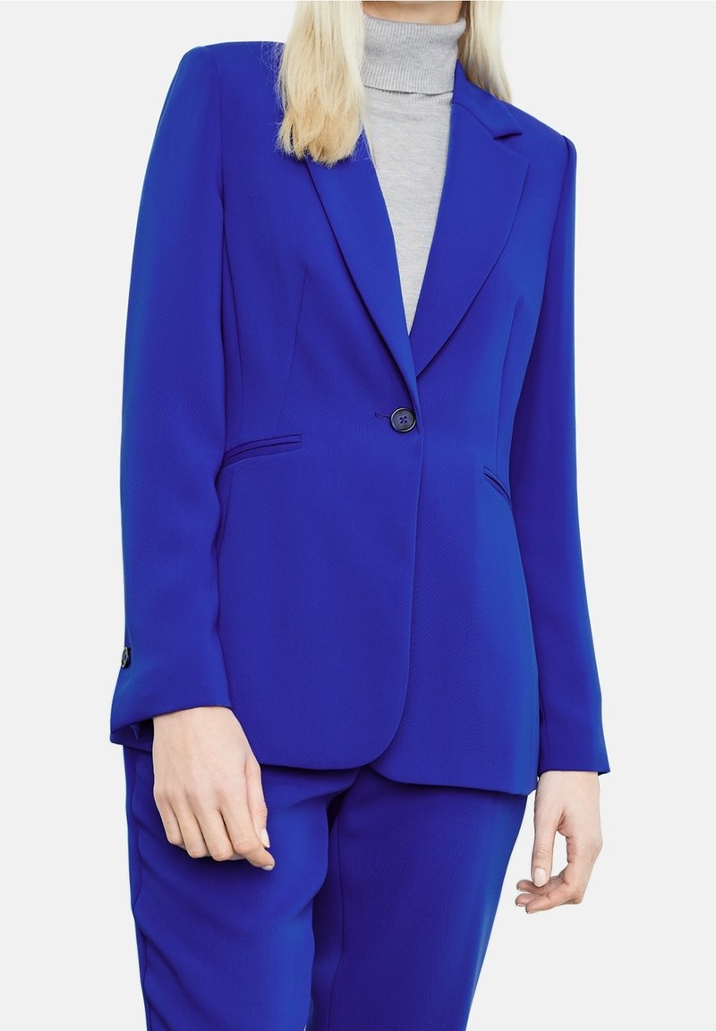 Femme portant un blazer bleu cobalt et un pantalon assorti sur un pull à col roulé gris clair, debout devant un fond blanc uni.