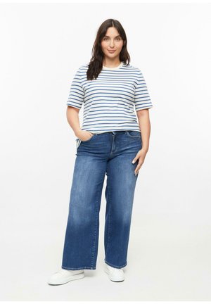 Femme aux cheveux foncés portant un t-shirt à manches courtes rayé blanc et bleu, un jean bleu à jambes larges et des baskets blanches, debout les mains dans les poches.