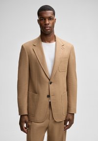 Maßgeschneiderter beiger Blazer mit weicher Textur, zwei aufgesetzten Taschen, schmalen Revers und zwei schwarzen Knöpfen, kombiniert mit einem weißen Rundhalsausschnitt-Shirt.