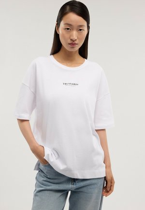 Joven con el cabello largo y oscuro que lleva una camiseta blanca de gran tamaño con el texto "Trussardi Gentle Society" y jeans azul claro contra un fondo liso.