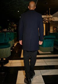 Abrigo de color azul marino oscuro con un corte a medida, visto desde atrás. Presenta un cuello alto, puños abotonados y pantalones y zapatos negros elegantes.