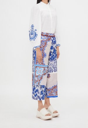 Personne portant un chemisier blanc avec broderie bleue sur les manches, un pantalon large à motifs bleu et marron, et des sandales blanches à plateforme.