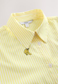 Geltonos ir baltos dryžuotos marškiniai, užsegami sagutėmis ir turintys apykaklę, šalia viršutinės sagos siuvinėtas mažas banano dizainas.