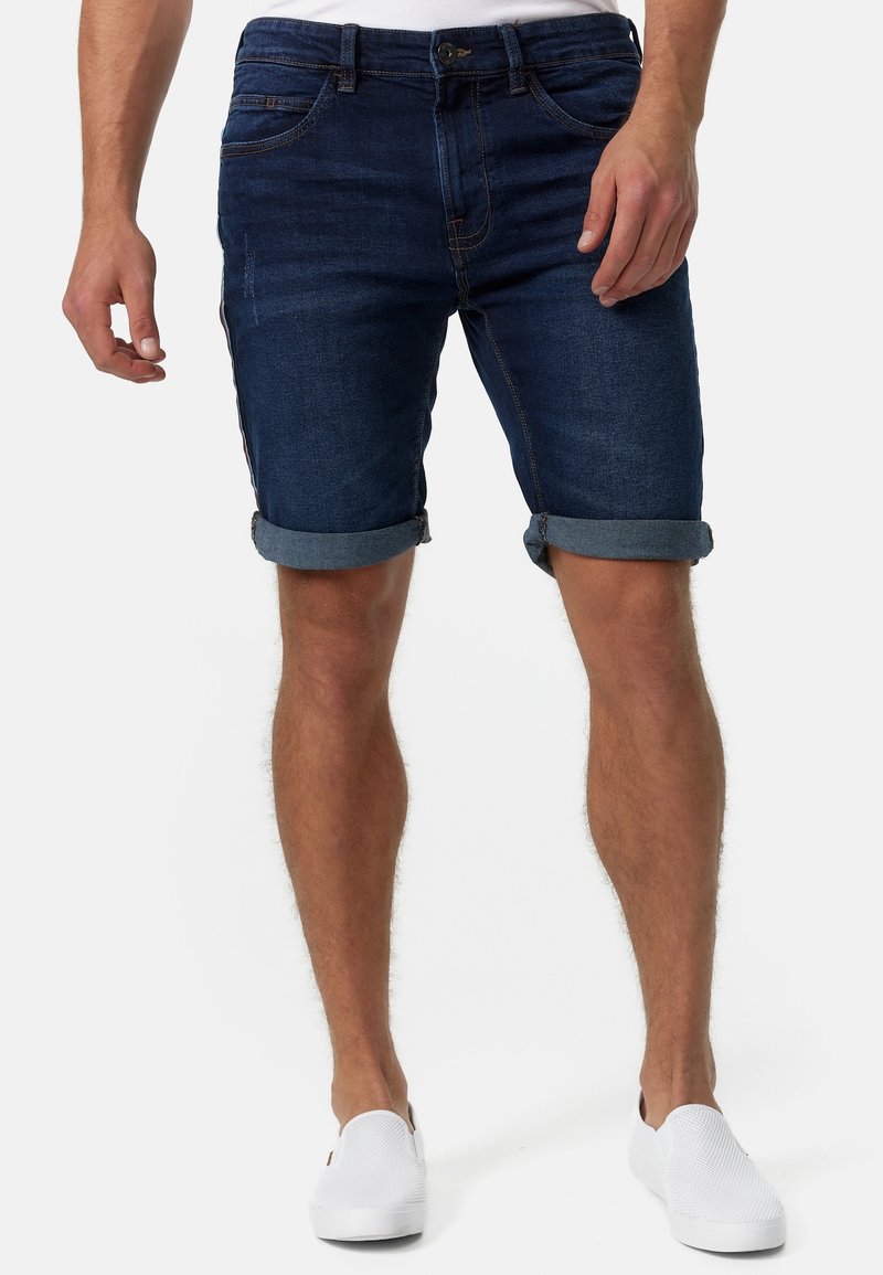 Homme vêtu d’un short en denim bleu foncé retroussé et de chaussures blanches sans lacets, debout les mains détendues le long du corps.