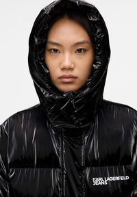 Junge Person mit schwarzen Haaren, die eine glänzende schwarze Steppjacke mit hochgestellter Kapuze trägt, das Karl Lagerfeld-Jeans-Logo ist auf der Brust sichtbar.