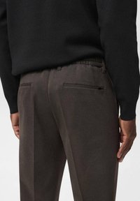 Pantaloni da uomo marrone scuro con una texture liscia, dotati di una cintura elastica e due tasche posteriori con cerniere a contrasto.