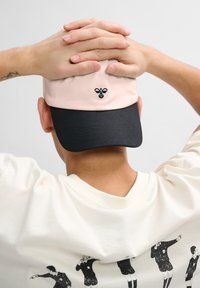 Cap with a pink crown and black visor, featuring a small embroidered bee logo. Worn backward with hands resting on top. White shirt visible.
