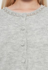 Gilet gris clair avec une texture douce, orné d'un col arrondi décoré de perles et de trois boutons en tissu sur le devant.