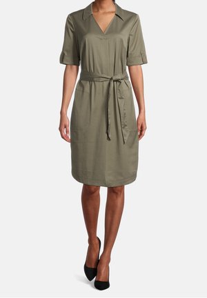 Kvinde iført knælængde olivengrøn kjole med korte ærmer, krave, forlommer og bindebælte i taljen, kombineret med sorte hæle.