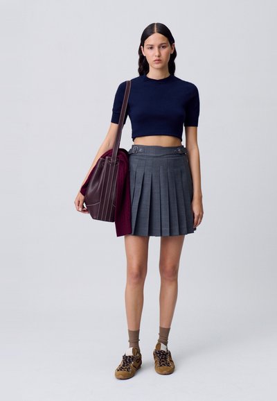 Top de punto recortado en azul marino, falda gris plisada con hebillas laterales, zapatillas marrones y bolso burdeos. La textura es suave con líneas estructuradas.