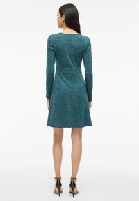 Teal long-sleeve dress with a fitted bodice and flared skirt. Textured pattern throughout, cinched at the waist, worn with black heels.