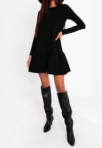 Czarna sukienka z długim rękawem i falbaną na dole, wykonana z miękkiego materiału. Noszona z czarnymi, wysokimi butami w stylu slouch, podkreślająca dopasowaną sylwetkę.