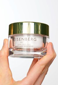 En klar glasburk som innehåller en vit kräm, toppad med ett glänsande guldlock. Texten på burken anger att det är en åldersbekämpande behandling från Eisenberg.