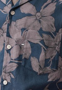 Blue textured fabric shirt with large gray floral print, white buttons, and a chest pocket.