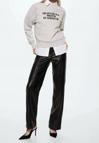 Felpa grigia con la scritta "QUELQUES MOTS D'AMOUR", indossata sopra a una camicia bianca, abbinata a pantaloni a gamba larga in ecopelle nera e tacchi neri.