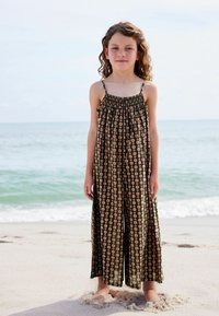 Blomstermønstret, sort ærmeløs jumpsuit med smocked liv og brede ben, båret af et barn, der står på stranden. Sand og hav i baggrunden.