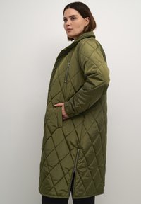 Jachetă lungă cu umplutură, de culoare verde măsliniu, cu un buzunar cu fermoar, buzunare laterale și o deschidere inferioară cu fermoar. Are o glugă și o textură netedă.