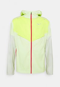 Chaqueta cortavientos Nike en verde claro y amarillo con cremallera frontal roja, logo Swoosh reflectante y capucha, mostrada sobre un fondo blanco.