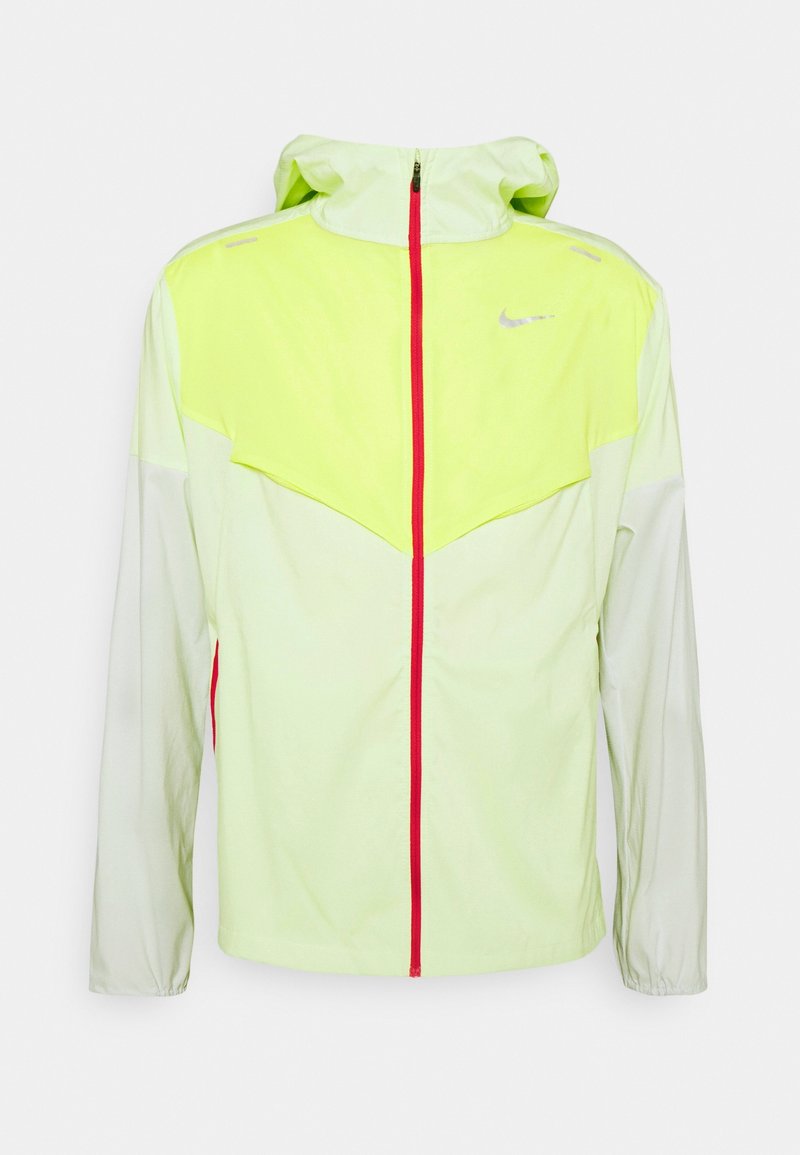 Chaqueta cortavientos Nike en verde claro y amarillo con cremallera frontal roja, logo Swoosh reflectante y capucha, mostrada sobre un fondo blanco.