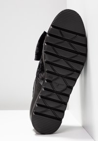Semelle de chaussure noire avec des rainures profondes et un motif texturé, inclinée contre un mur et un sol blancs.