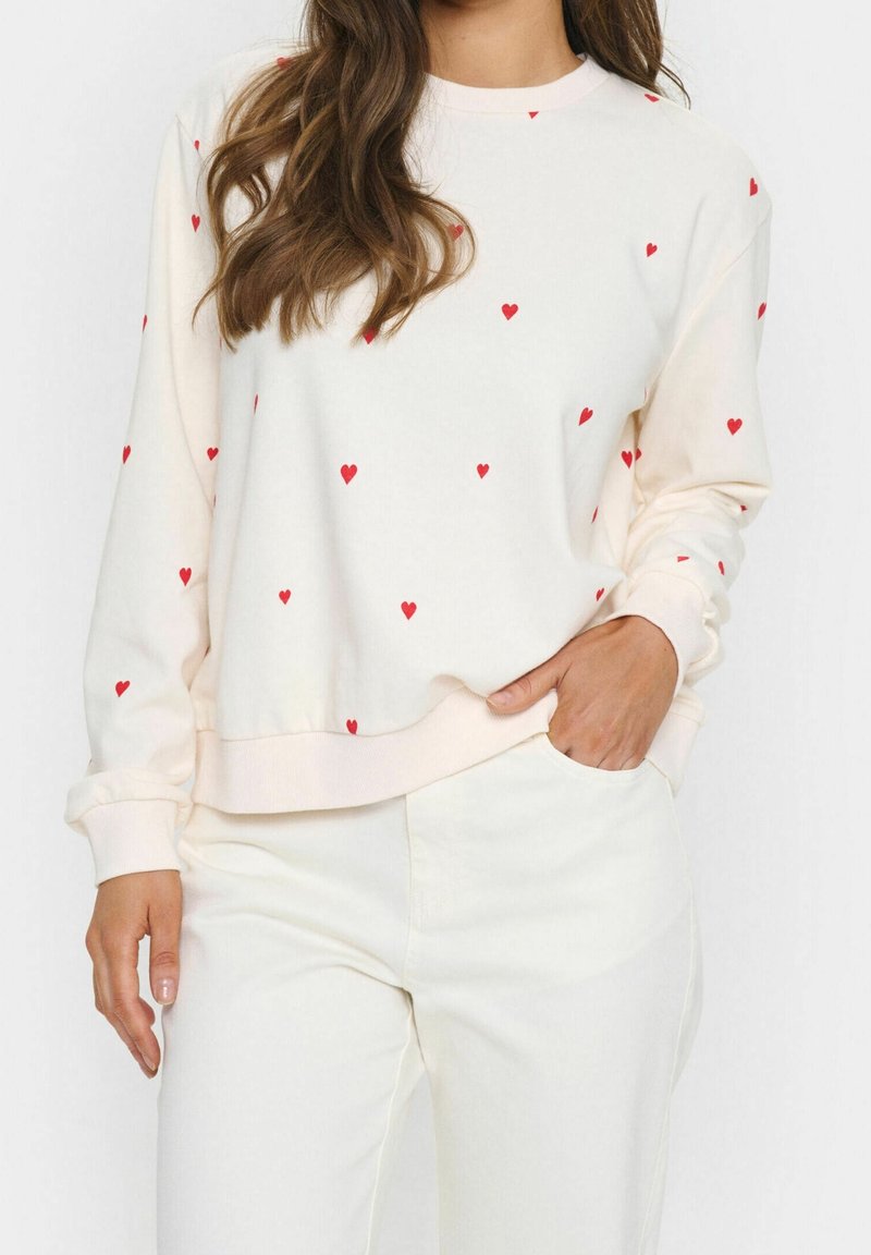 Femme portant un sweat-shirt écru avec de petits cœurs rouges et un pantalon écru assorti, main posée négligemment dans la poche.