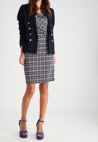 Blazer blu navy con bottoni argentati sopra un abito a motivo bianco e nero. La modella indossa tacchi blu con plateau e posa su uno sfondo chiaro.