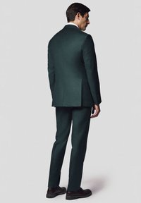 Ciemnozielony garnitur z dopasowanym krojem, z jednym rozcięciem z tyłu oraz białą koszulą elegancką. Czarne buty dopełniają całość. Materiał ma gładką fakturę.