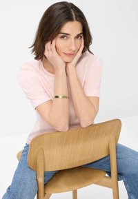 Top rosa chiaro a maniche corte con scollo rotondo, abbinato a jeans blu. Il modello è seduto su una sedia di legno, mostrando un bracciale d'oro sul braccio.